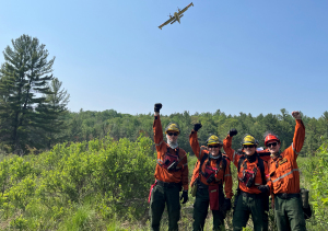 A group of four FireRangers in Personal Protective Equipment stand together with an arm raised in the sky. A waterbomber aircraft flies through the sky behind them.