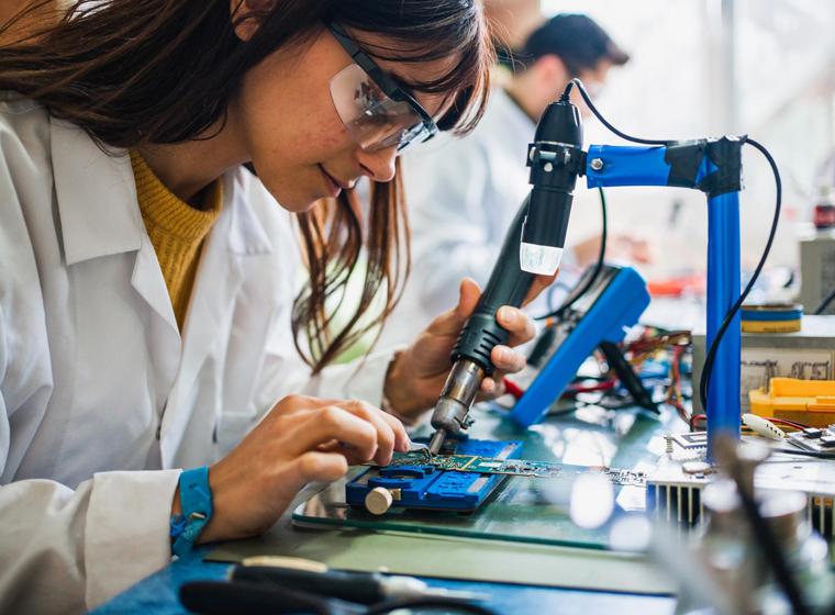 A student wearing a white lab coat is using a soldering iron to work on an electronic circuit board.