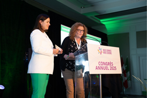 The Honorable Natalia Kusendova‑Bashta, Minister of Long‑Term Care, presents the visual identifier to Barbara Ceccarelli, Executive Director of the Centre d’accueil Héritage, in front of a podium displaying a sign indicating the name of the Assembly of the Francophonie’s 2025 annual conference..