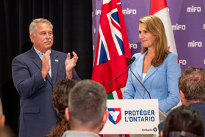 L’Honorable Caroline Mulroney, ministre des Affaires francophones, prend la parole à la tribune, avec les drapeaux de l’Ontario et du Canada en arrière‑plan ainsi qu’un panneau affichant le logo du MIFO, tandis que l’Honorable Neil Lumsden, ministre du Sport, applaudit lors d’une annonce publique.