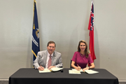 Photograph of Ontario’s Minister of Francophone Affairs, the Honourable Caroline Mulroney, wearing a pink dress, and Billy Nungesser, Lieutenant Governor of Louisiana, wearing a grey suit, seated side by side at a table while holding a signed joint declaration, with the Ontario and Louisiana flags displayed in the background.