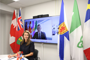 Photograph ofntario’s Minister of Francophone Affairs, The Honorable Caroline Mulroney, wearing a black dress, seated in front of a white table while signing a joint statement. Behind her, the Minister of Acadian Affairs and Francophonie of the Government of Nova Scotia, Colton LeBlanc, appears on a screen. Beside the screen are the flags of Canada, Ontario, Franco‑Ontarian, Nova Scotia, and Acadia.
