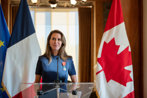 The Honorable Caroline Mulroney, Minister of Francophone Affairs, speaking at the podium, with the French and Canadian flags in the background, during her induction into the Legion of Honour.