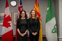 Photograph of Ontario’s Minister of Francophone Affairs, the Honourable Caroline Mulroney, and Assistant Deputy Minister Maria Lamani standing side by side, both wearing black attire, with the flags of Canada, Ontario, Franco‑Ontarian, Acadia, and New Brunswick displayed in the background.