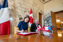 Photograph of Ontario’s Minister of Francophone Affairs, the Honourable Caroline Mulroney, and the Ambassador of France to Canada, Michel Miraillet, seated side by side at a wooden table while both sign a joint declaration. Small flags are displayed on the table, with the Canadian and French flags in the background.