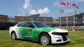 A vehicle of the Ontario Provincial Police, displaying green insignia representing Franco‑Ontarian francophonie, is parked in front of a public building. Five flags are visible in front of the building: the Franco‑Ontarian flag, the Ontario flag, the Canadian flag, the Ontario Provincial Police flag, and the 2ELGBTQI+ flag.